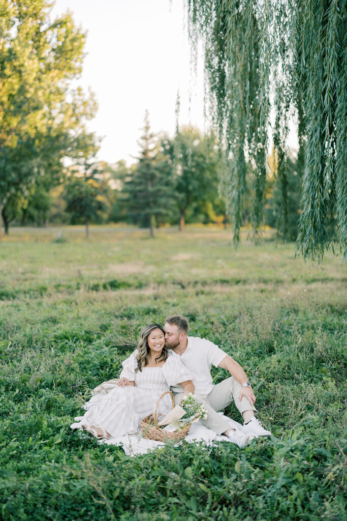 engagement session, a couple sitting in a green field on a picnic blanket under a willow tree